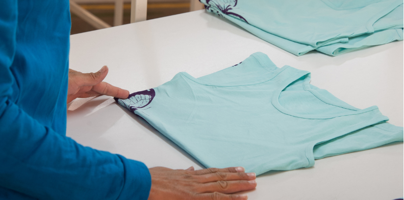 A person folding a light blue t-shirt on a table, wearing a blue long-sleeved shirt. The t-shirt has a small design on the sleeve, and the person is gently pressing down on the fabric to fold it neatly.
