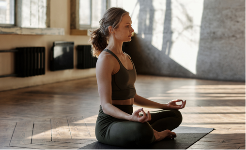 The image shows a person sitting cross-legged on a yoga mat in a well-lit room with large windows and wooden floors. The person is wearing a dark sports bra and dark leggings, and is in a meditative pose with hands resting on the knees, palms facing upward, and fingers forming a mudra. The room has a serene and calm atmosphere, with sunlight streaming in and casting shadows on the floor.