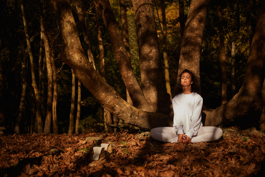 A woman practicing yoga meditation outdoors in a fall forest, wearing white yoga attire.
