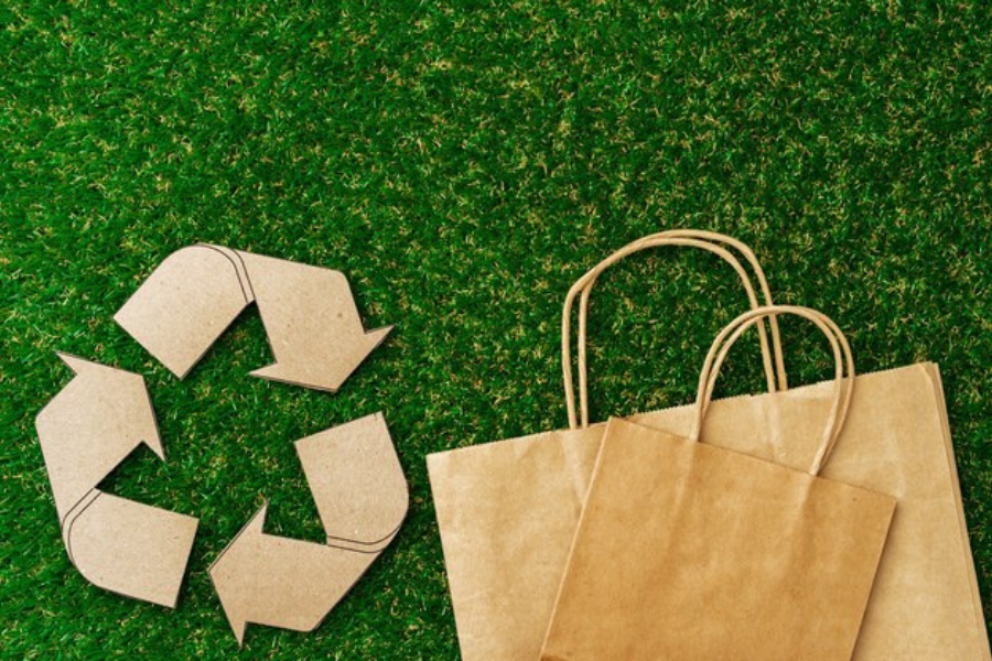 Recycling symbol made from cardboard on green grass, with eco-friendly brown paper bags beside it, representing sustainable practices in packaging