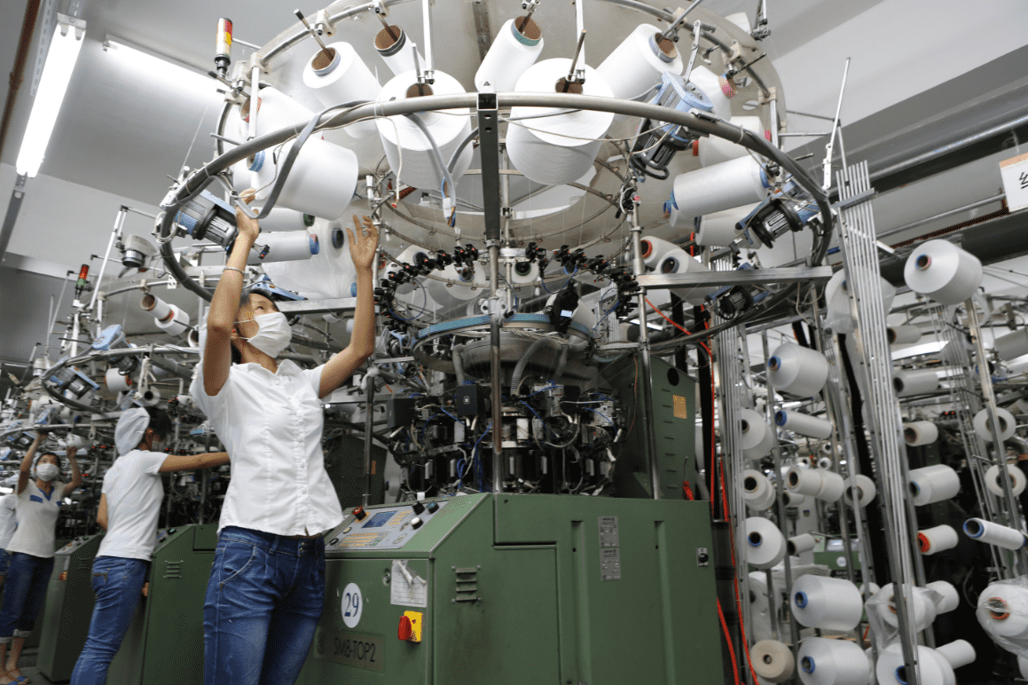 The image shows workers in a textile factory operating large textile machines. The workers are wearing white tops and jeans, adjusting and checking the spools of yarn on the machines. Numerous spools of yarn surround the machines, illustrating the busy and complex nature of textile production.