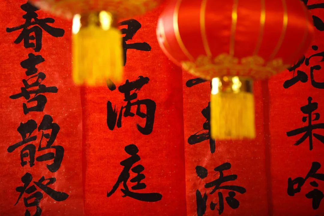The image shows red Chinese lanterns and red banners with black calligraphy. The lanterns are adorned with golden tassels. The banners contain vertical Chinese characters, commonly used as decorations during celebrations such as the Lunar New Year. The text on the banners likely conveys auspicious blessings and wishes for happiness, prosperity, and good fortune.