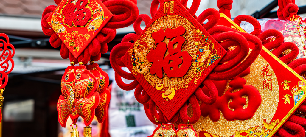 The image shows traditional Chinese New Year decorations. The decorations are predominantly red and gold, symbolizing good fortune and prosperity in Chinese culture. The main elements include a large red diamond-shaped decoration with the Chinese character "福" (fu), which means "blessing" or "good fortune." This decoration is adorned with intricate designs, including flowers and gold accents. Below it hangs a pair of red and gold fish decorations, symbolizing abundance and wealth. Another red and gold decoration with the same "福" character is surrounded by floral patterns and additional Chinese characters likely conveying wishes for prosperity and happiness.