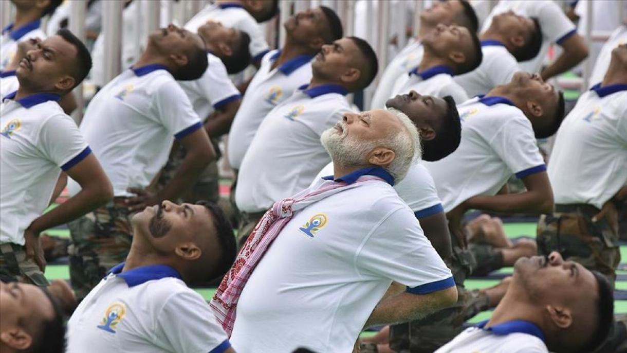 The image shows a group of people performing a yoga pose together, including Indian Prime Minister Narendra Modi. They are all wearing white shirts with blue collars and a logo on the left side of the chest, which appears to be related to yoga. The individuals are bending backward with their hands on their hips and looking upwards. This organized event seems to be a yoga session or class with multiple participants performing the same pose in unison, emphasizing collective physical activity and unity through yoga.